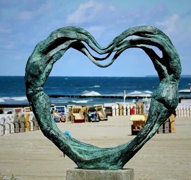 Rzeźba w kształcie serca na plaży, z morzem i niebem w tle.